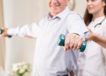 Retiree training with dumbbells assisted by young physiotherapist