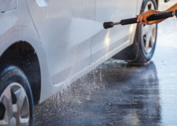 hand with high pressure washer washing white car at public self-service car washing station, close-up with selective focus