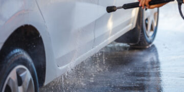 hand with high pressure washer washing white car at public self-service car washing station, close-up with selective focus