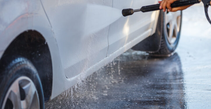 hand with high pressure washer washing white car at public self-service car washing station, close-up with selective focus